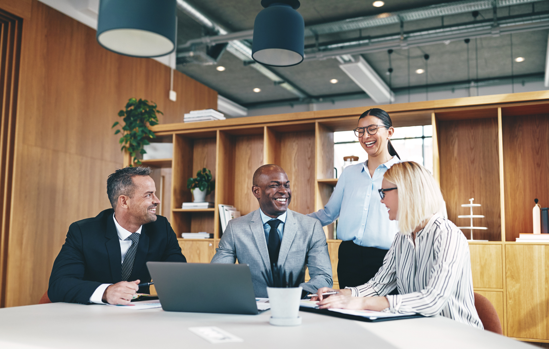 Four businesspeople in a meeting, smiling and looking at a laptop, in a modern office.