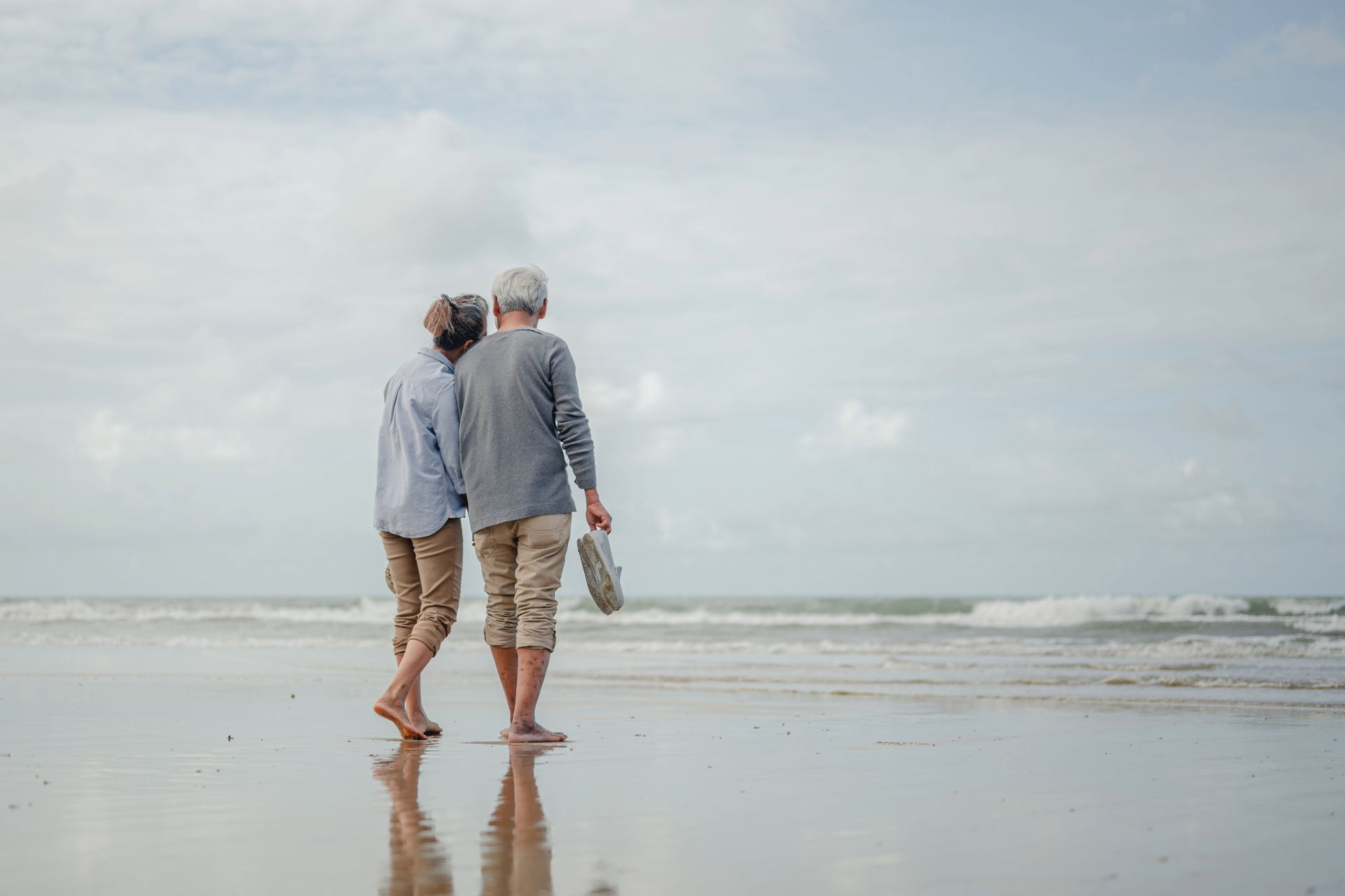 Senior couple walking barefoot on a beach, holding hands and gazing at the ocean under a cloudy sky.