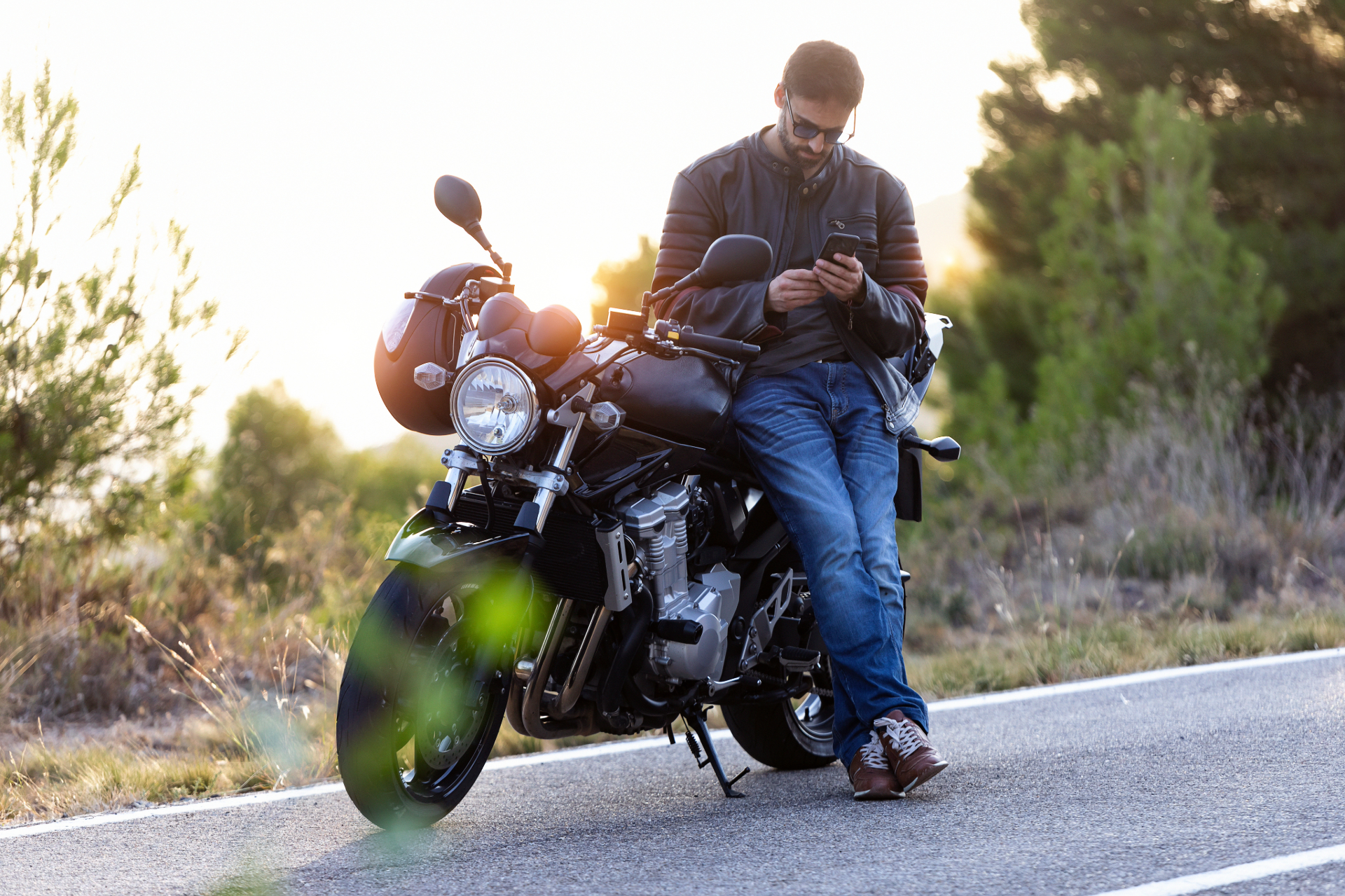 Man leaning against a motorcycle, using his phone, outdoors with sunset.