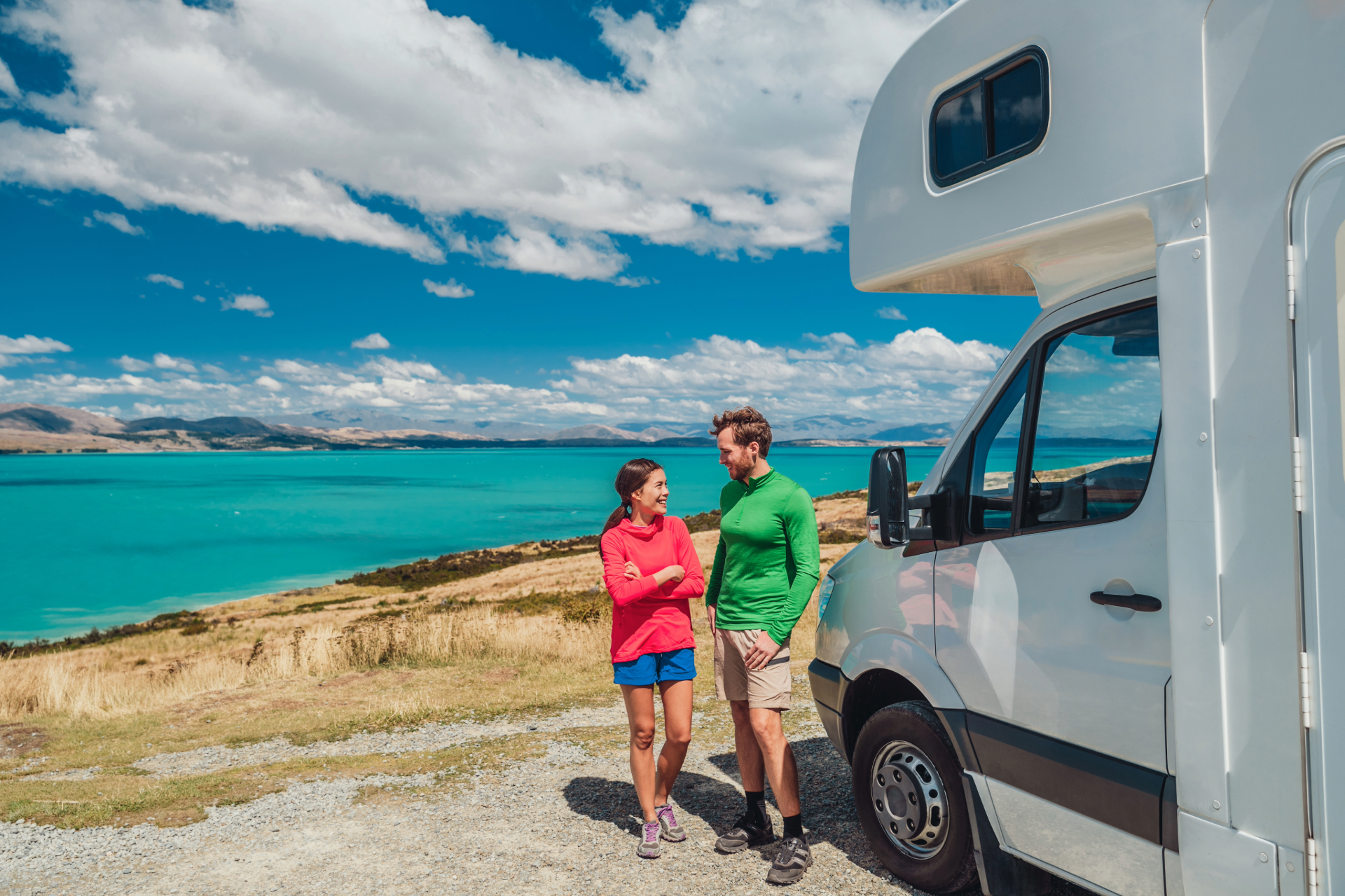 Couple standing near RV, overlooking turquoise lake and mountains under a blue sky.