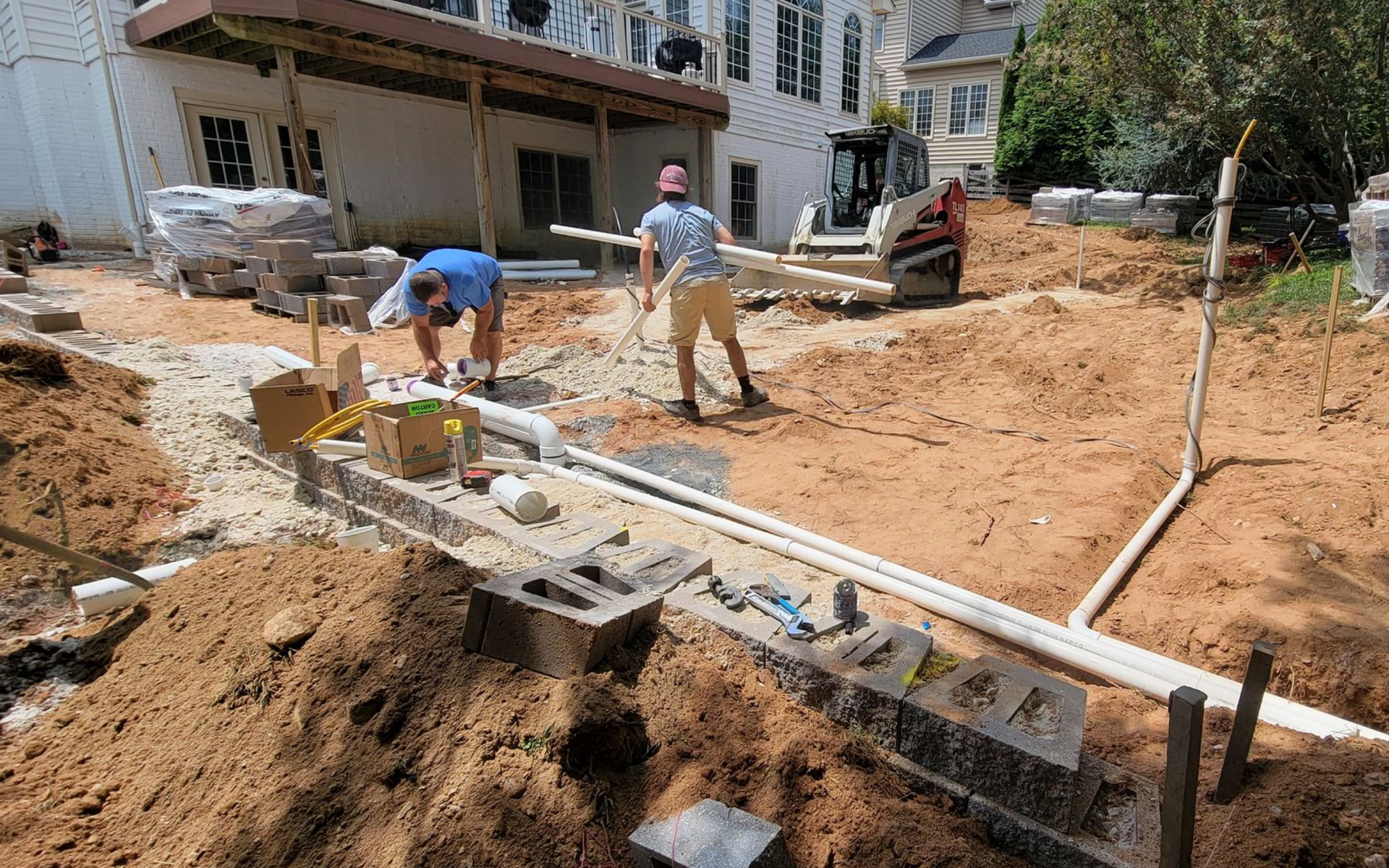 Construction workers building an outdoor patio. One cuts something while another carries lumber, with machinery and materials nearby.