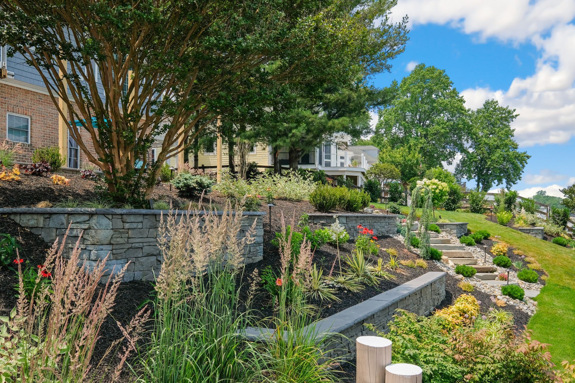 Terraced garden with stone retaining walls, flowering plants, trees, and a house in the background