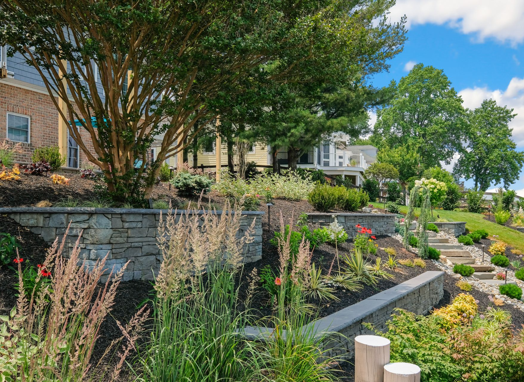 Landscaped garden with tiered stone retaining walls, plants, and trees under a blue sky.