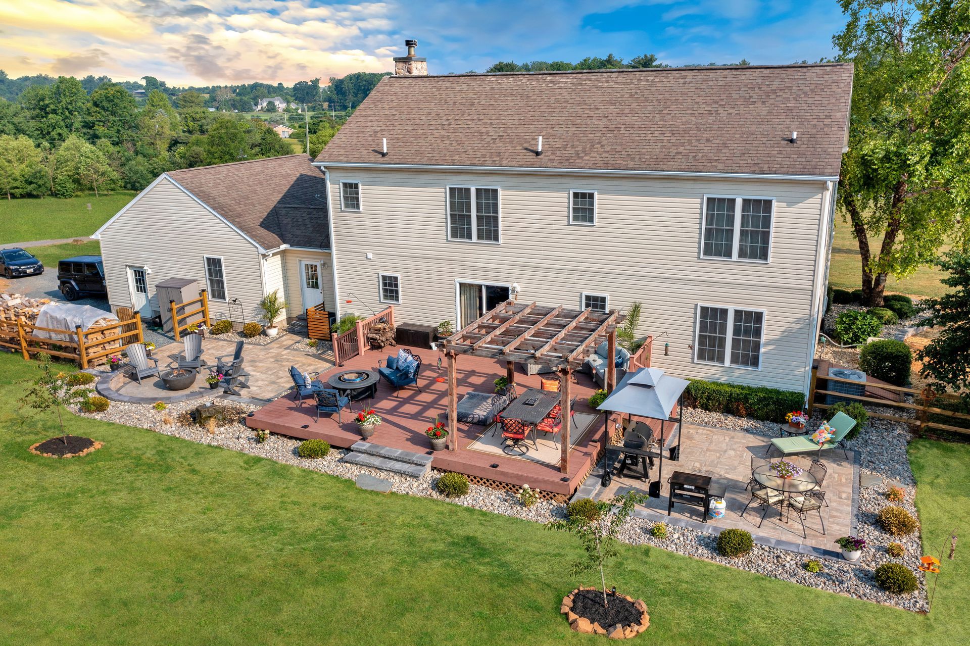 Backyard view of a two-story beige house with a wooden deck and patio, surrounded by green grass and landscaping.