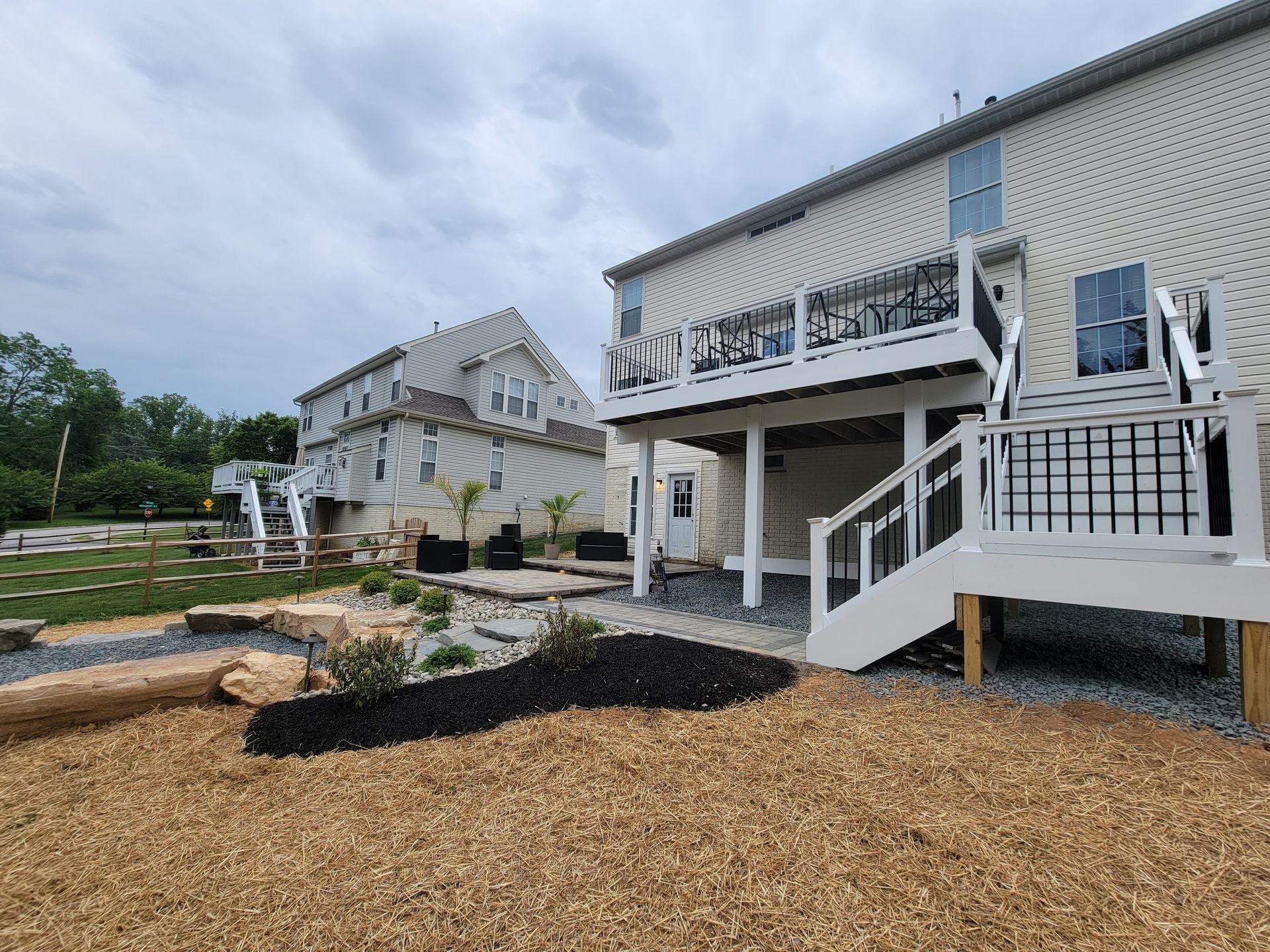 A backyard with a raised white deck, gravel ground covering, mulch landscaping beds, and a view of a neighboring house.