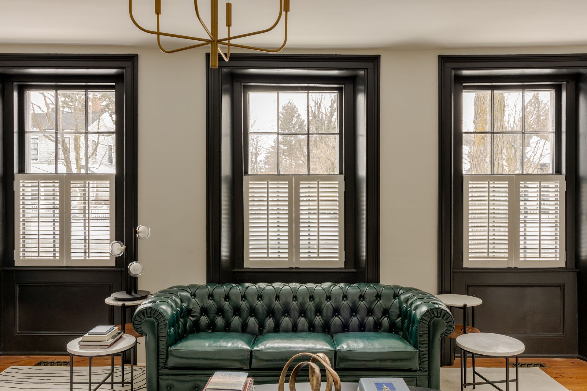 A tufted green leather Chesterfield sofa sits centered in a room with three large windows framed by dark, bold molding.