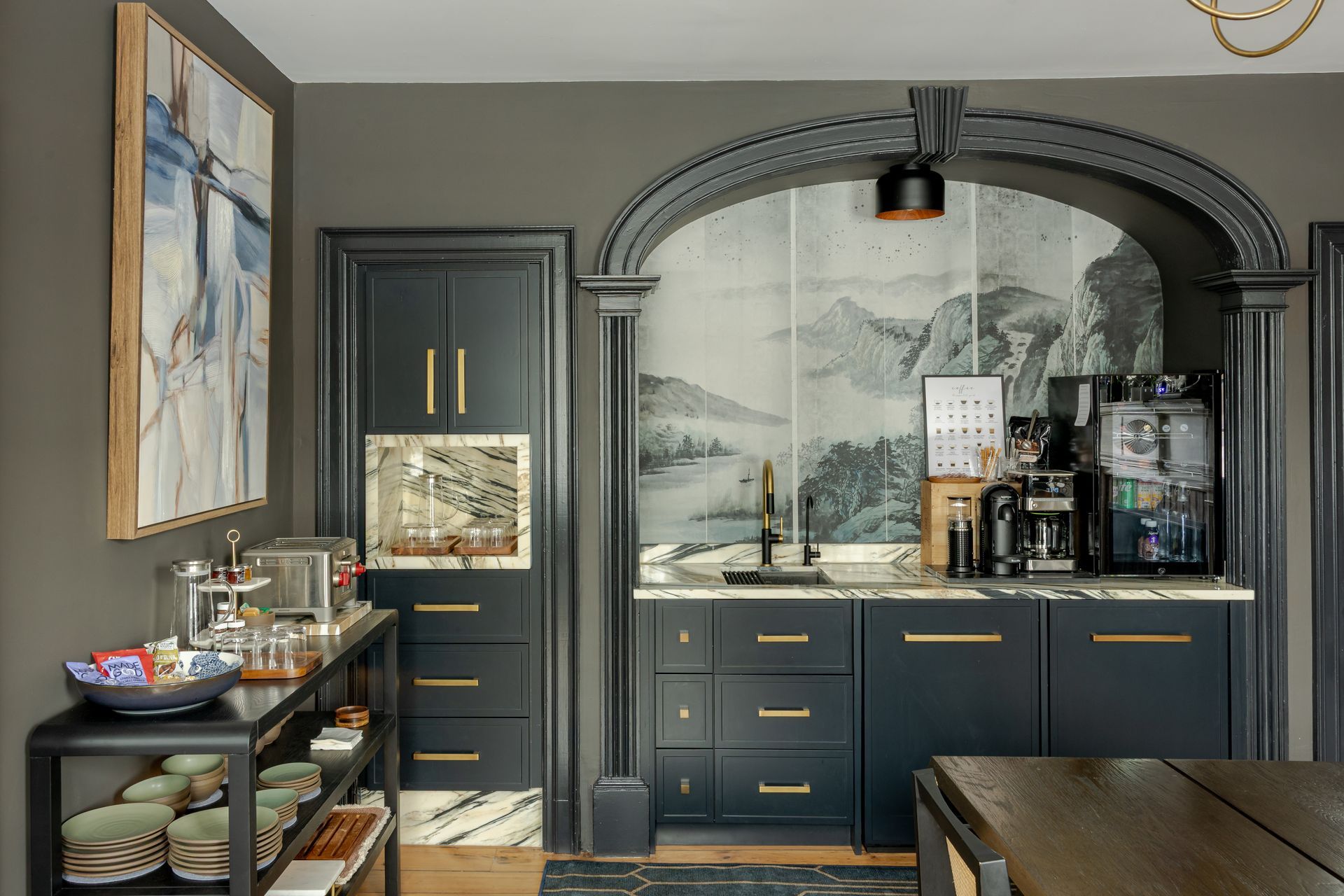 A dark-toned coffee bar with gold hardware, set within an arched alcove featuring mural wallpaper and a marble counter.