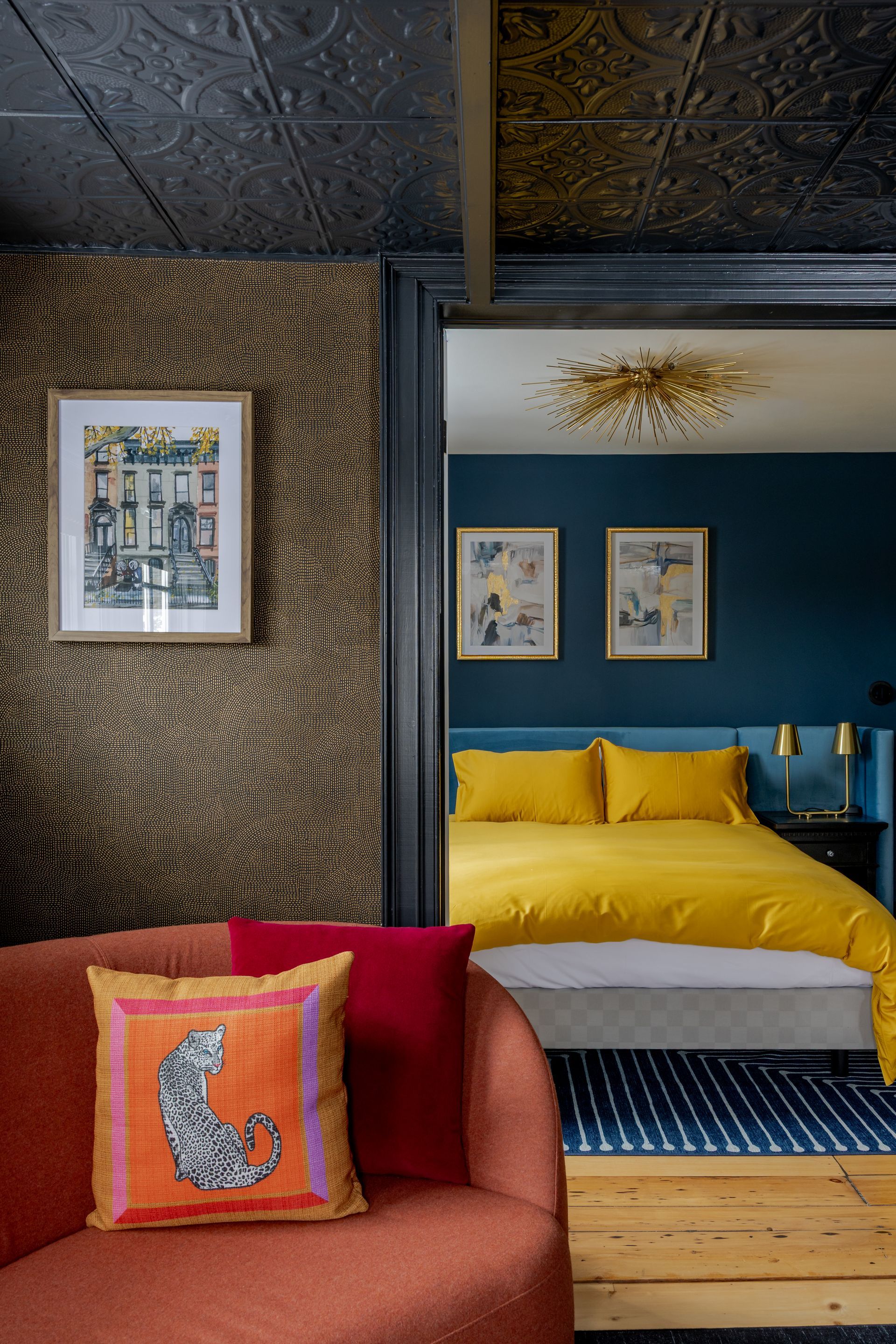 A burnt orange sofa with a patterned pillow faces an open doorway leading to a bedroom with mustard bedding and blue walls.