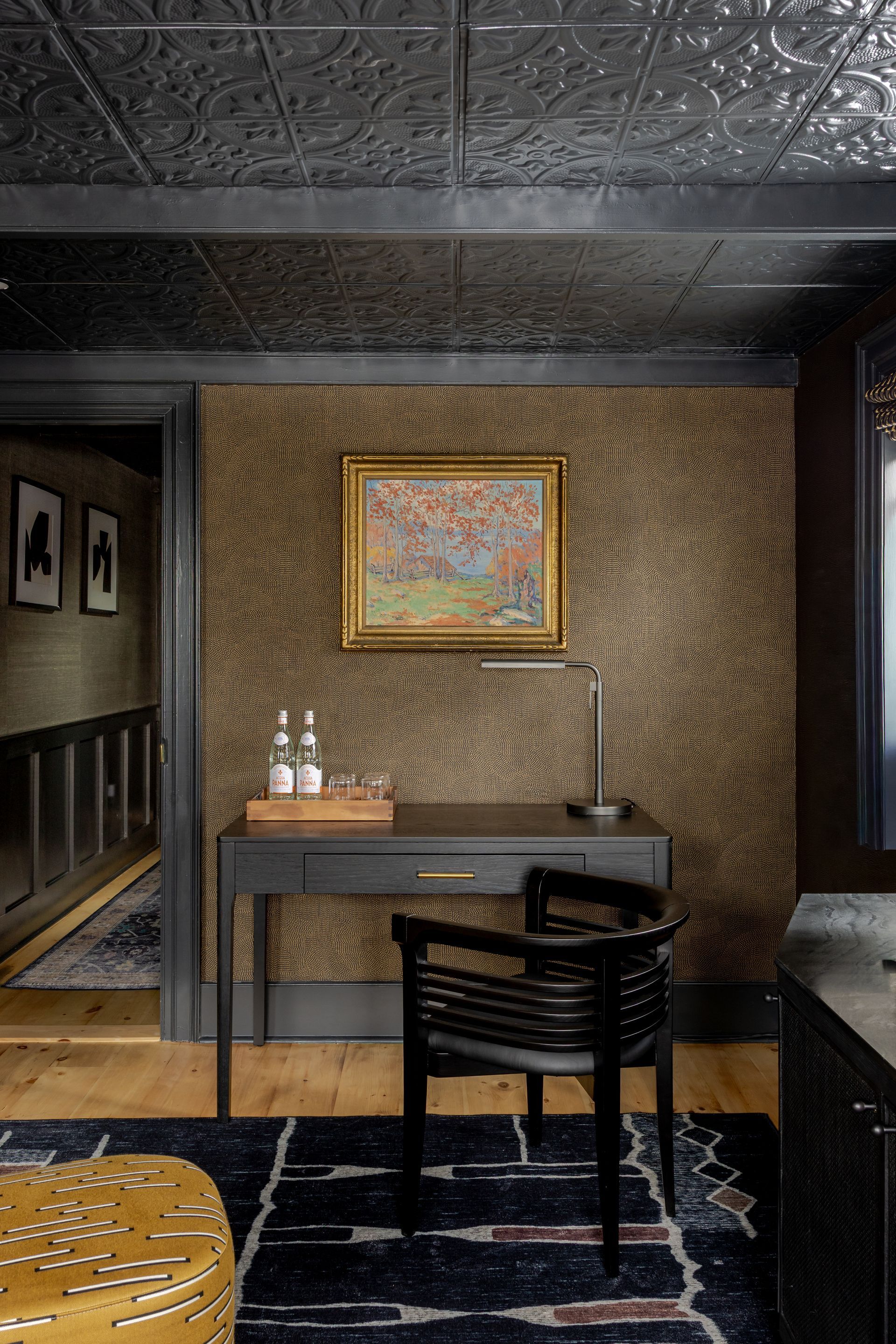 A dark desk and black chair sit against a textured brown wall below a framed painting, set on a dark, patterned rug.