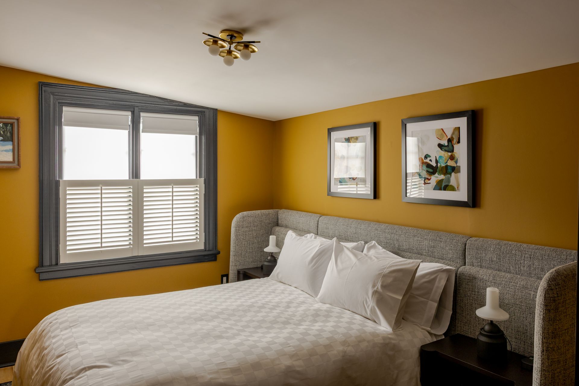 A cozy bedroom featuring mustard-yellow walls, a gray upholstered bed frame, white bedding, and a window with white shutters.