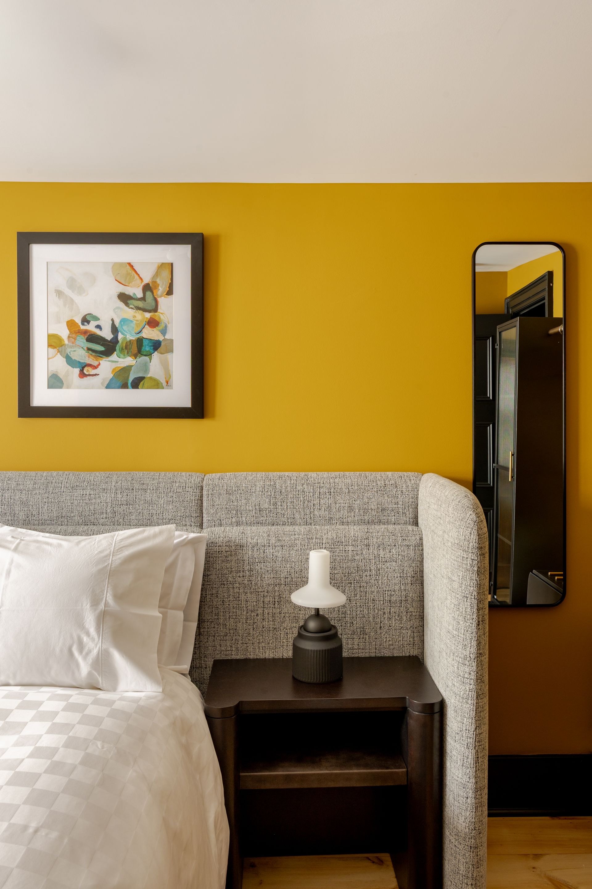 A mustard yellow wall with a framed abstract print, a black wall mirror, and a textured headboard with a small lamp.