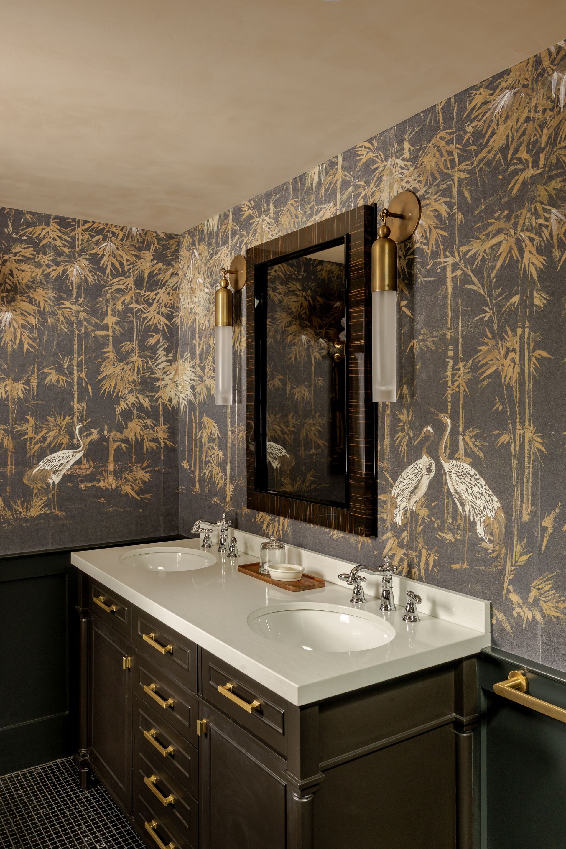 A bathroom vanity with a white countertop and dark cabinetry against wall-paper featuring gold trees on a dark background.