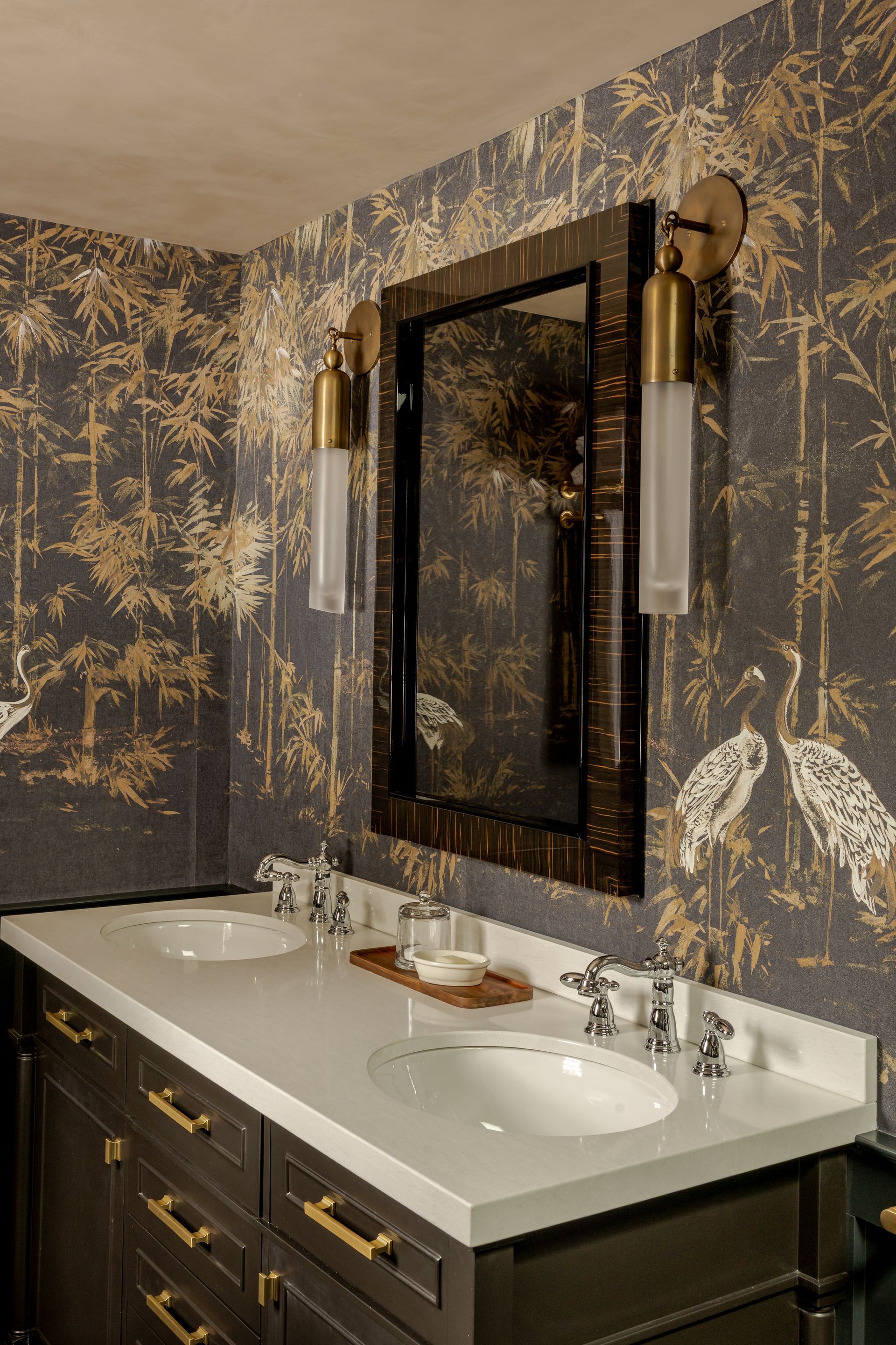 A bathroom vanity with a white countertop, two sinks, and dark cabinetry against wall-paper with a gold crane pattern.
