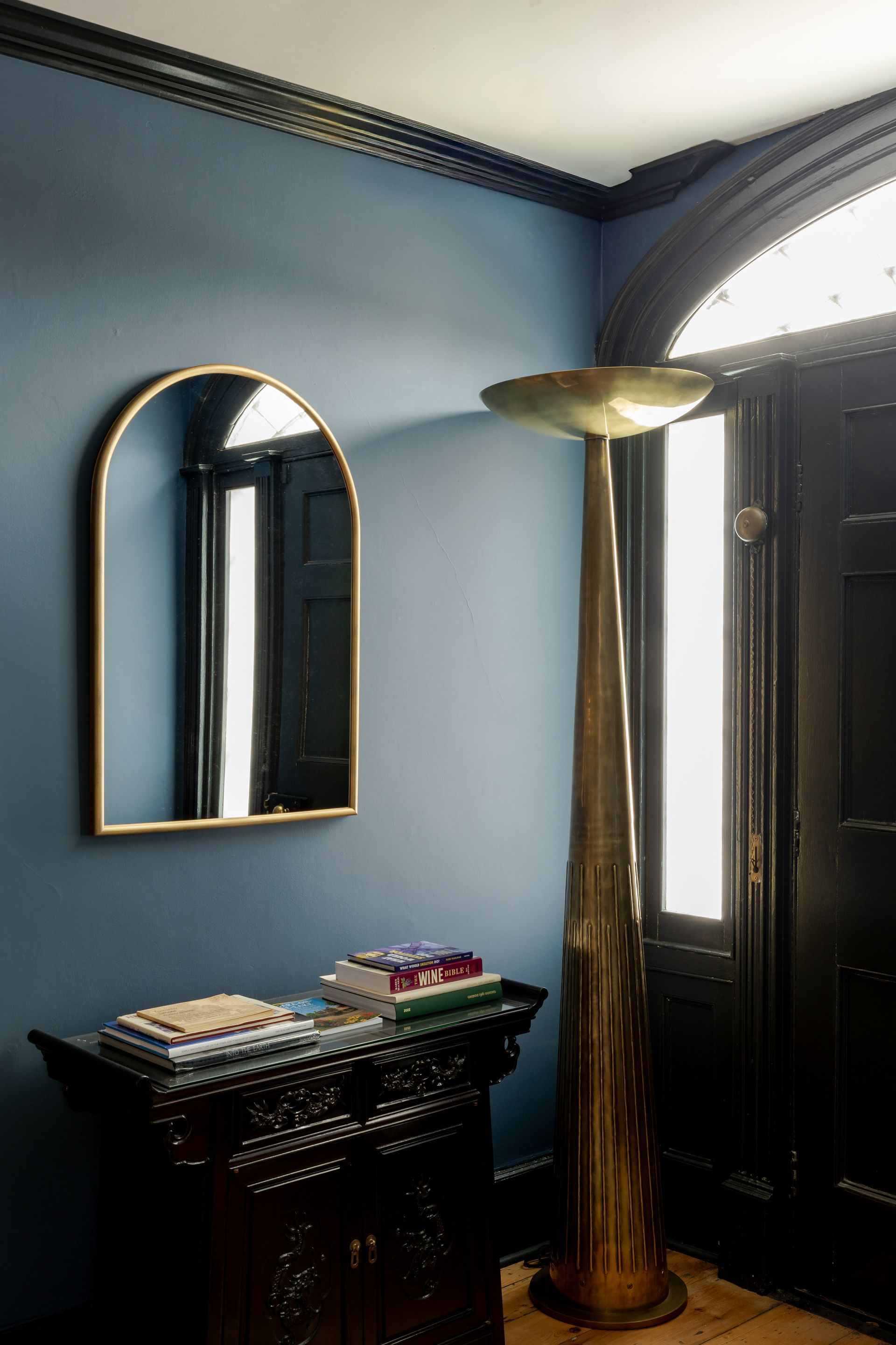 A foyer with blue walls, an arched gold mirror, a dark wooden console table with items, and a tall gold floor lamp.
