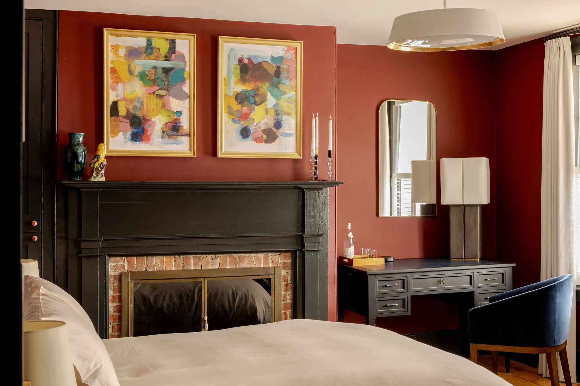 A bedroom with deep red walls, a black fireplace mantle, two abstract paintings, a vanity desk, and a bed in the foreground.