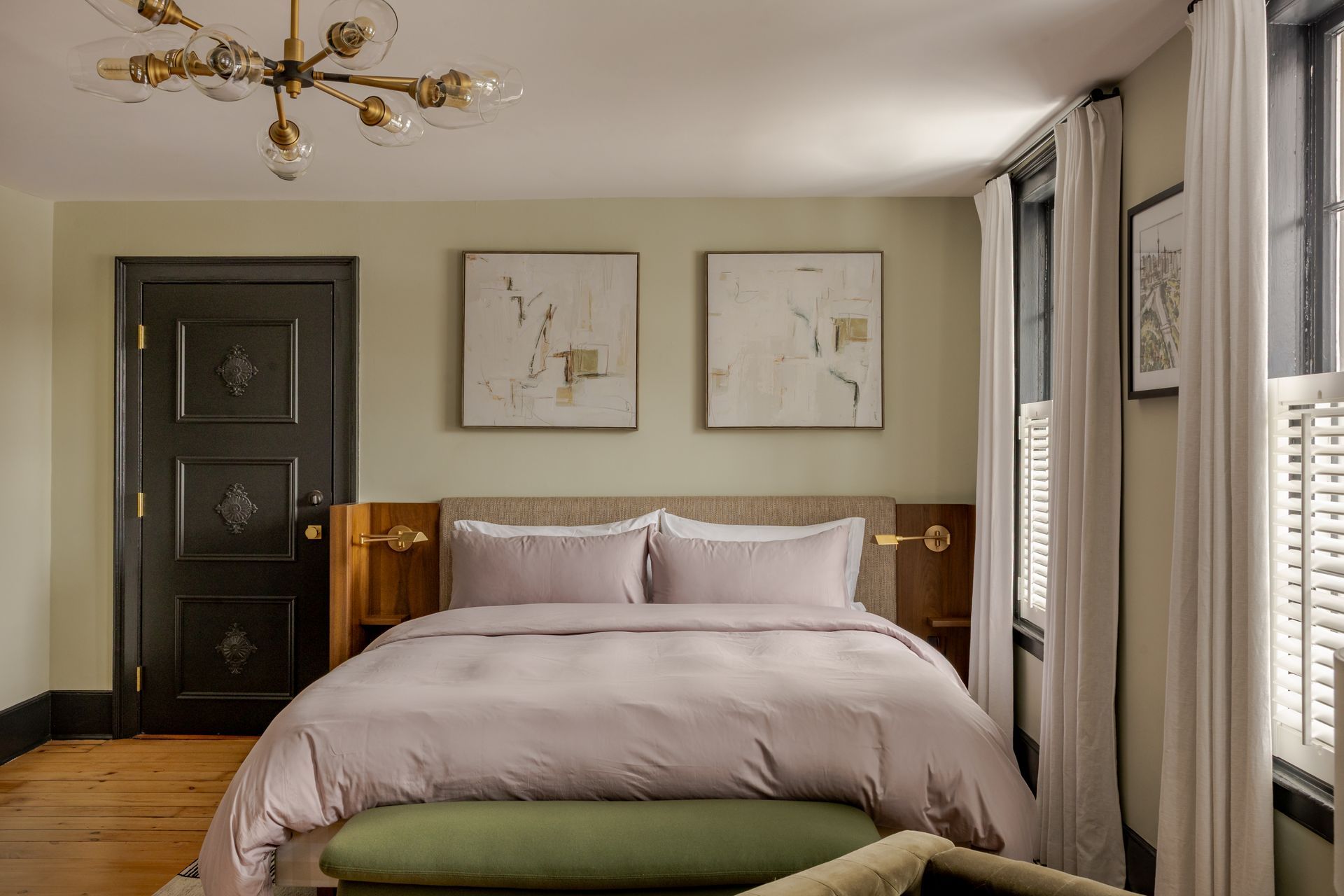 A cozy bedroom featuring a queen bed with mauve bedding, a sage green bench, two abstract wall prints, and a chandelier.
