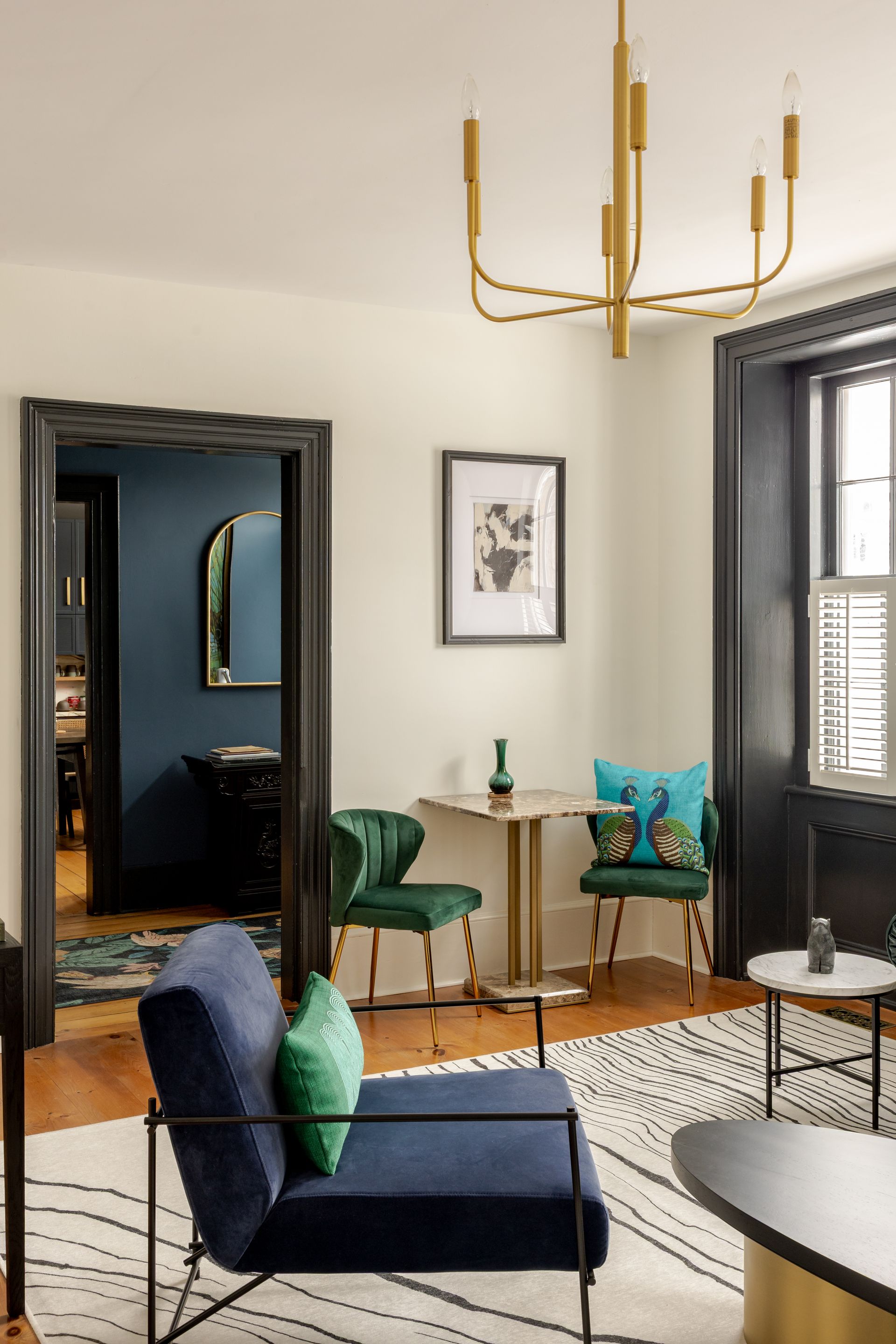 A modern living room with a navy armchair, two green velvet chairs, a pedestal table, and a black-and-white patterned rug.