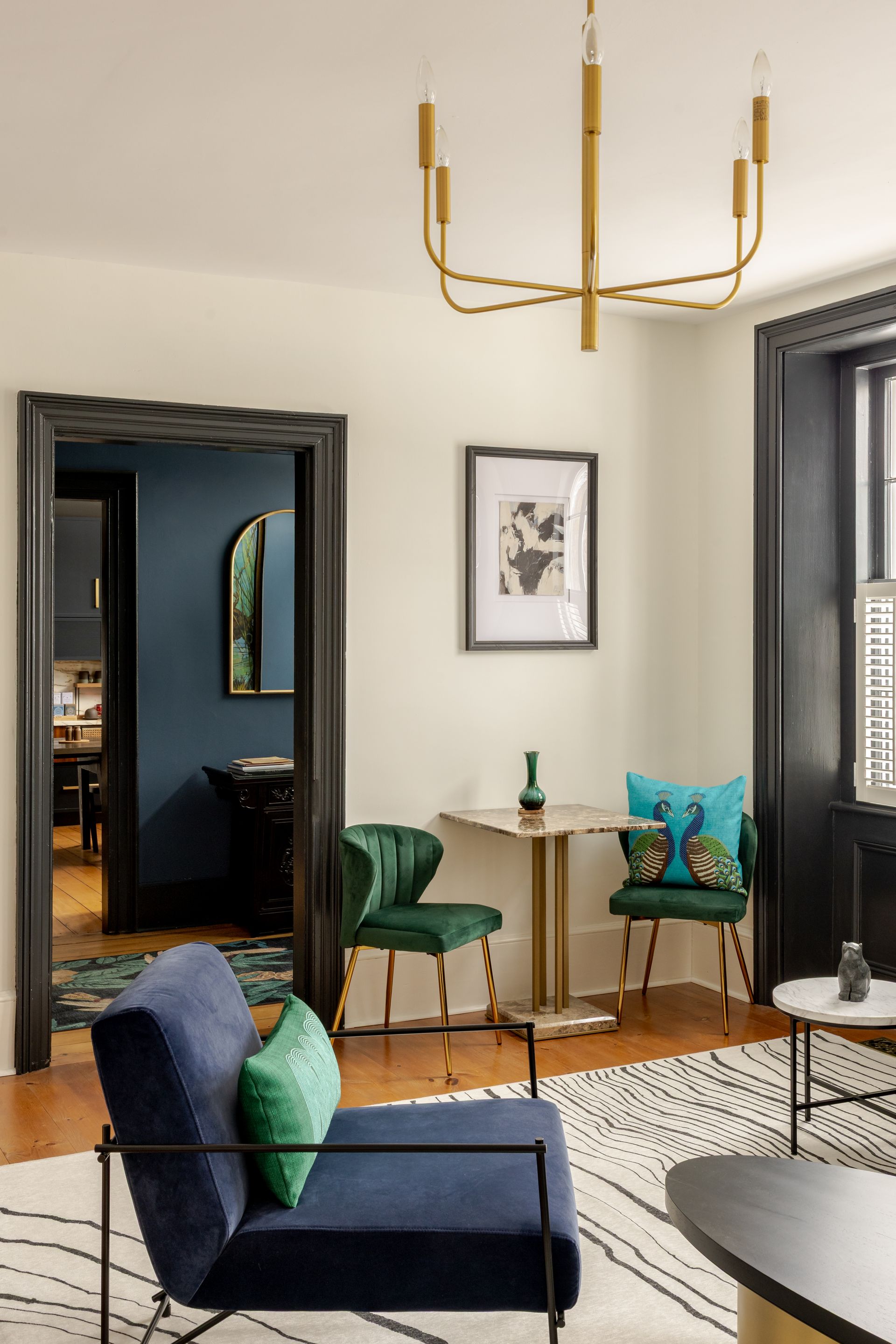A modern living room with a blue armchair, a small dining table with green chairs, and a gold chandelier on white walls.