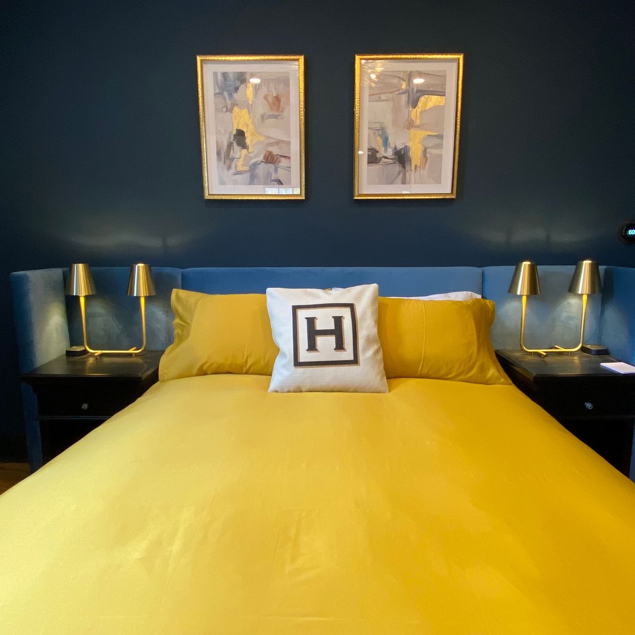 Bedroom with yellow bedding, blue headboard, nightstands, gold lamps, and artwork.