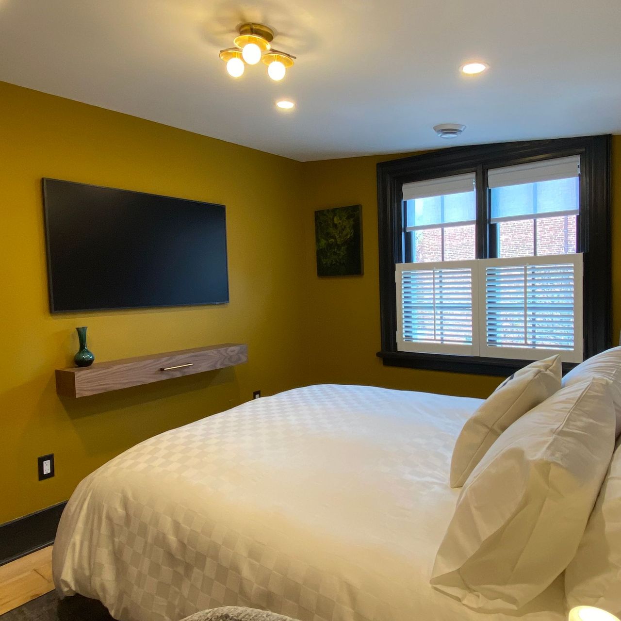 Bedroom with a bed, TV on wall, and window with blinds. Yellow walls, black window frame.