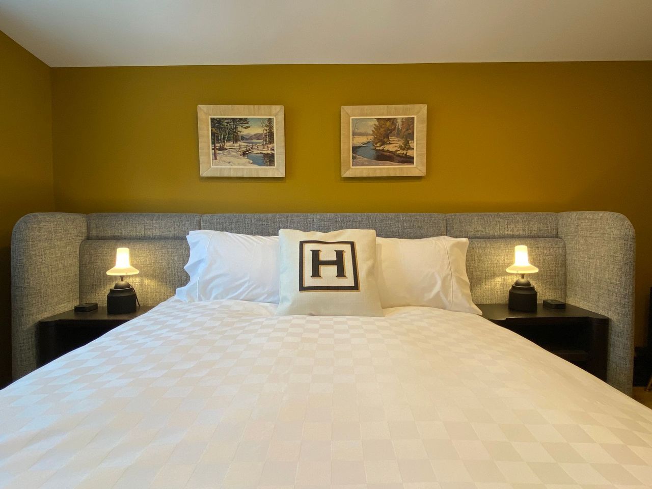 Bedroom with large bed, mustard-colored wall, two framed pictures, bedside lamps, and a decorative pillow.
