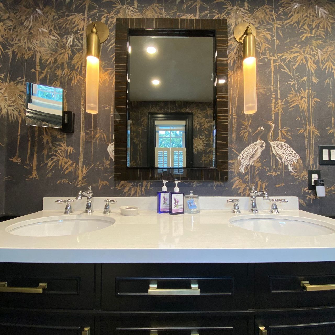 Bathroom with black vanity, white countertop, double sinks, patterned wallpaper, and gold sconces.