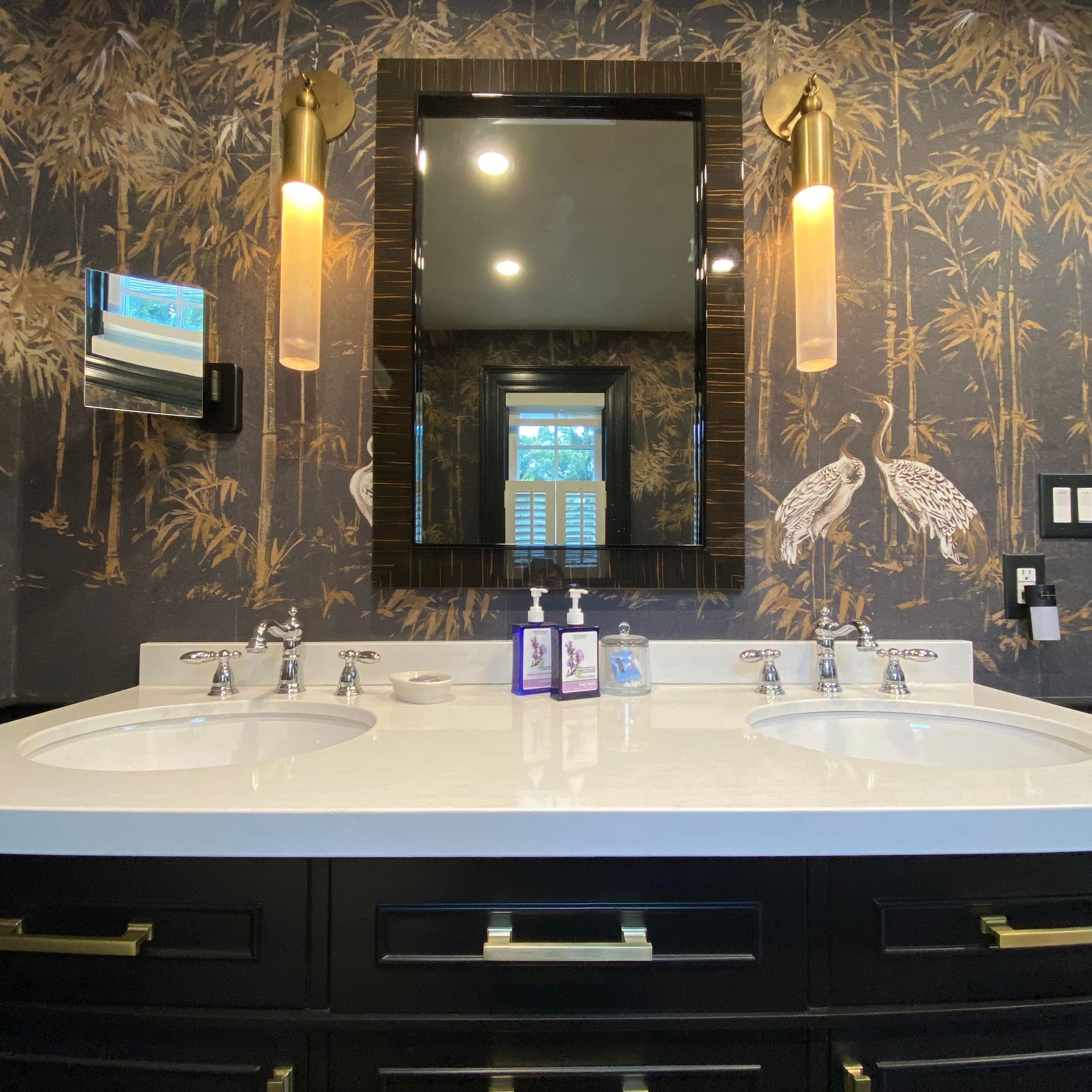 Bathroom with black vanity, white countertop, double sinks, patterned wallpaper, and gold sconces.