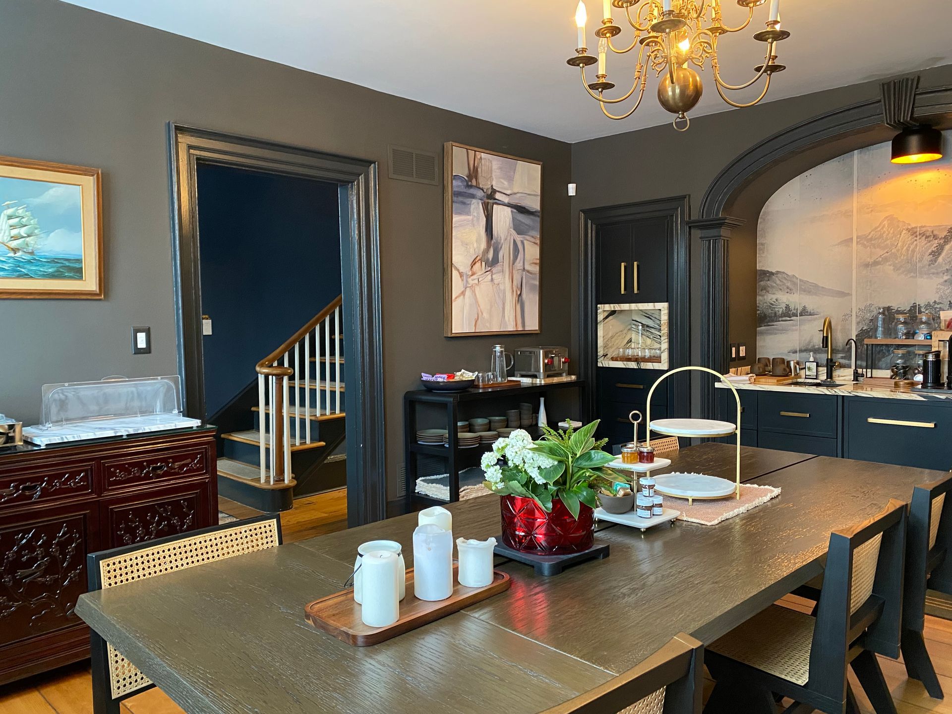 Dining room with gold table, dark walls, stairs, art, and chandelier.