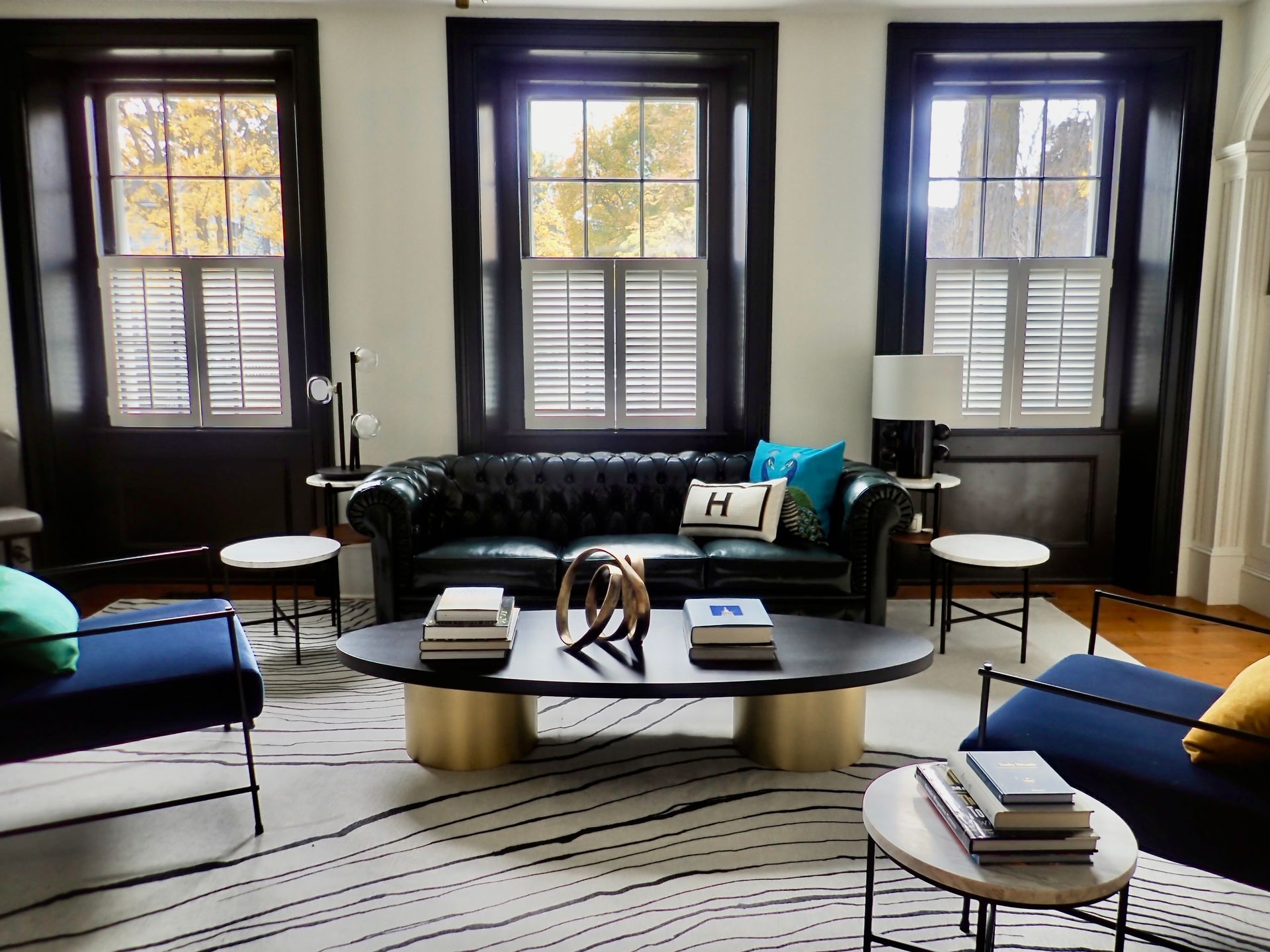 Living room with black leather sofa, chairs, and coffee table, three windows with shutters, and patterned rug.