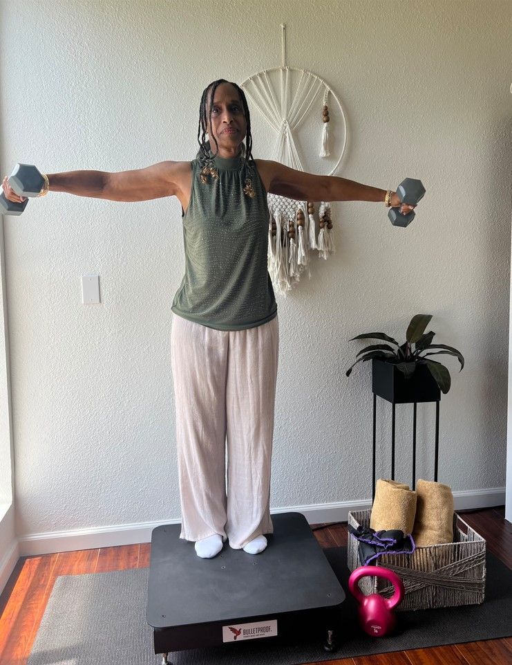 A woman is standing on a vibration plate holding dumbbells in her hands.