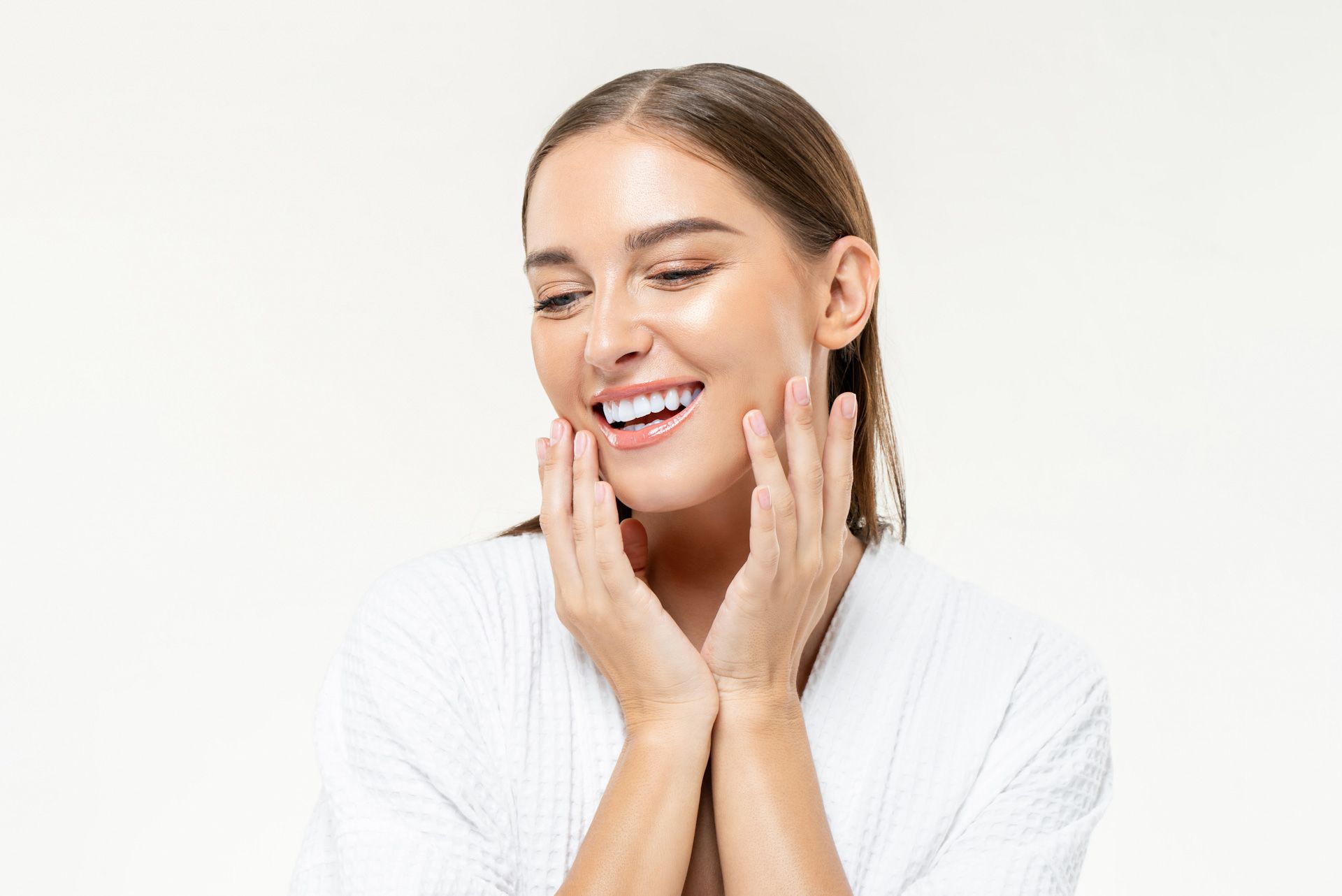 A woman in a white robe is smiling and touching her face.