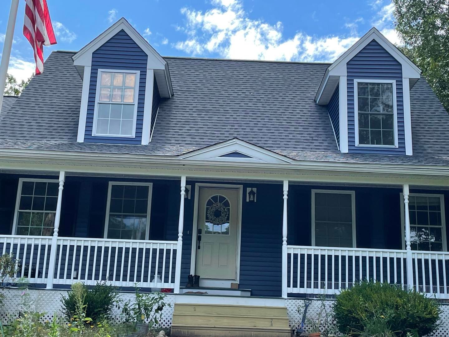 Blue house with white trim, porch, windows, and dormers; American flag in the background.