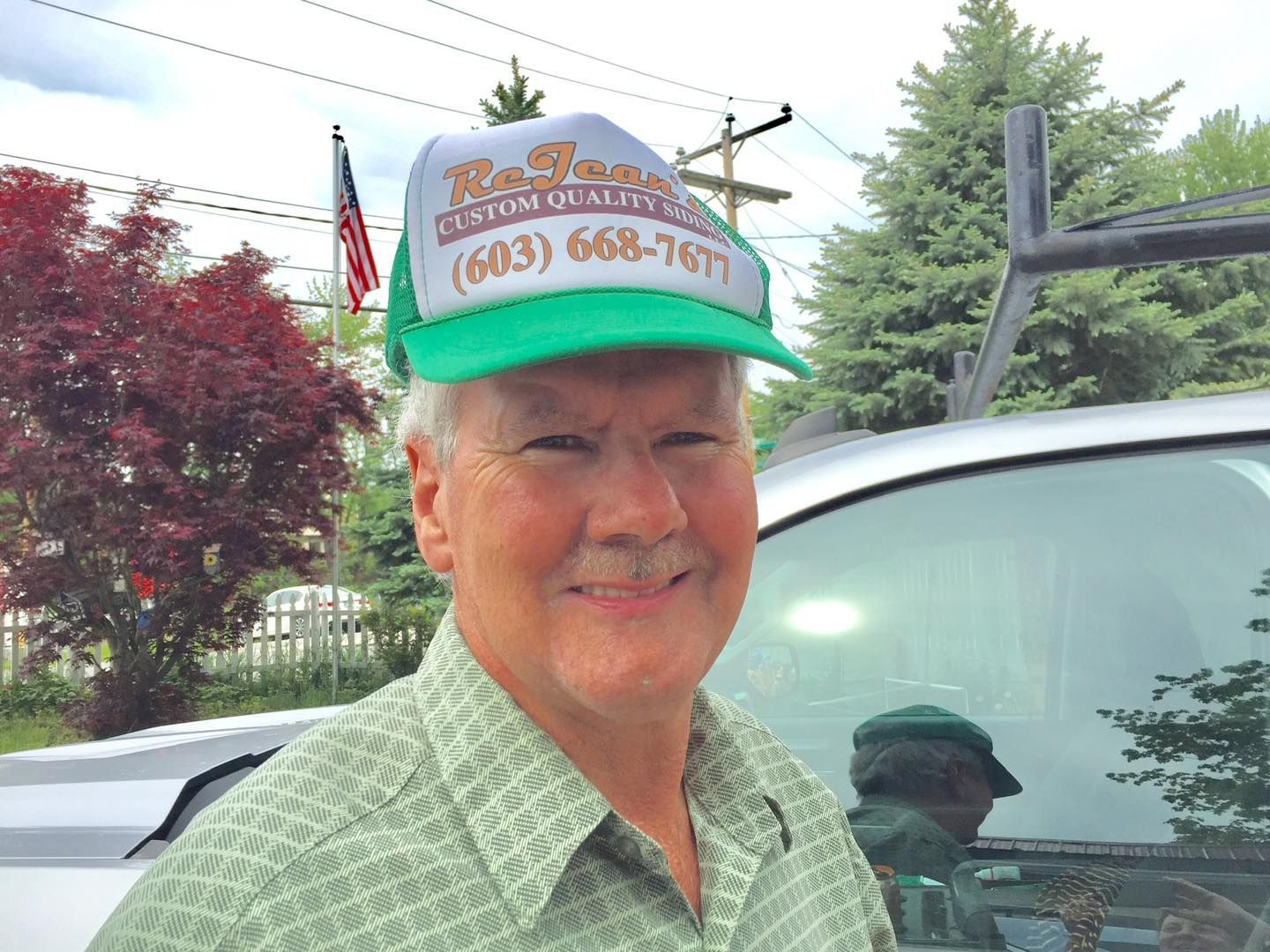 Man wearing green cap and shirt smiles outside, beside a window and American flag in background.