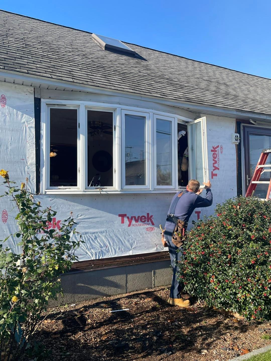 Man installing window on a house exterior. The house is covered in Tyvek. Bright blue sky.