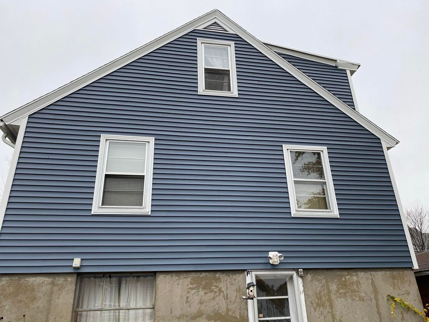 Blue-sided house with white-framed windows and a small door on a concrete foundation, against an overcast sky.