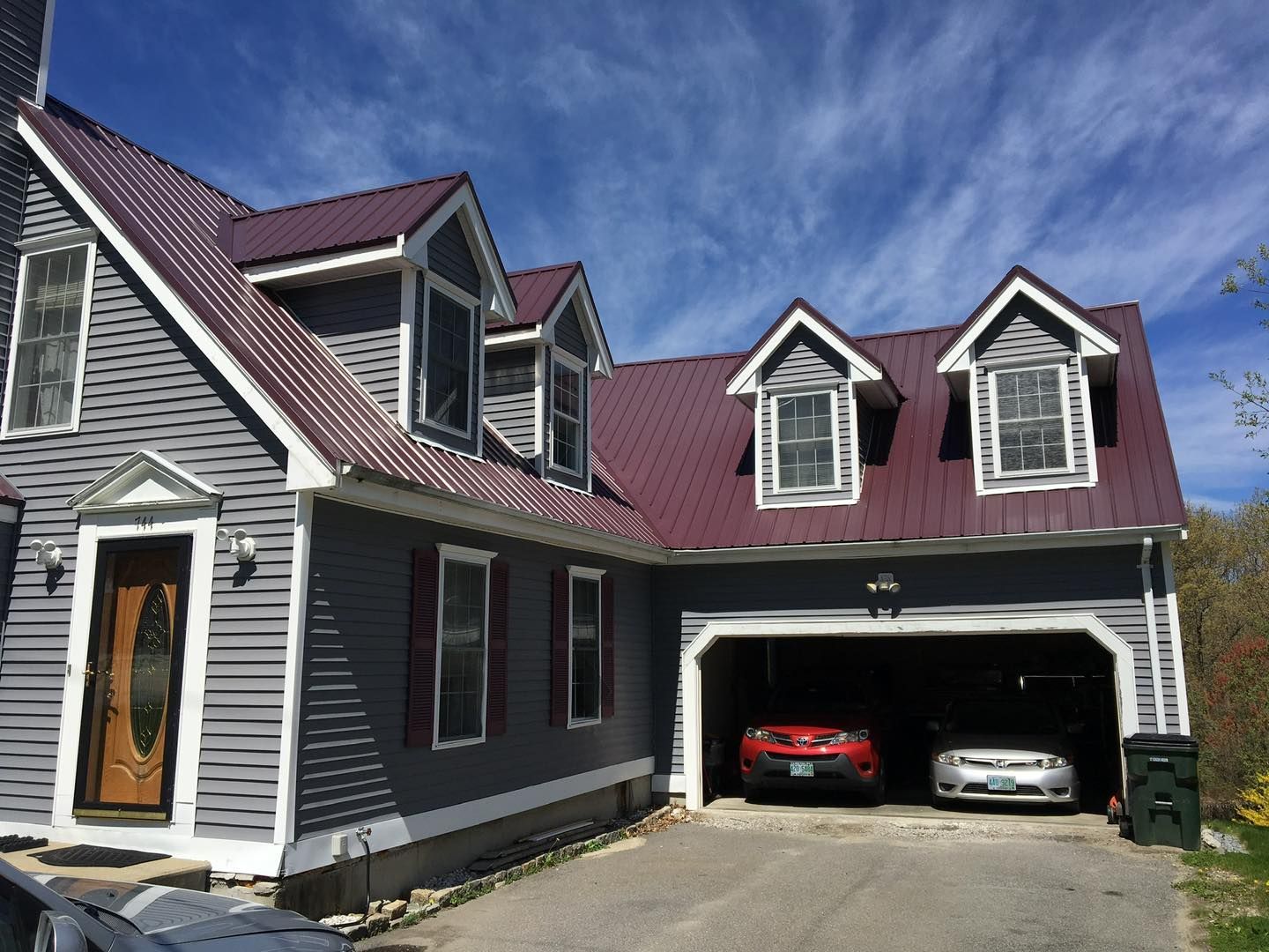 Gray house with burgundy metal roof, two-car garage, two cars inside, and blue sky.