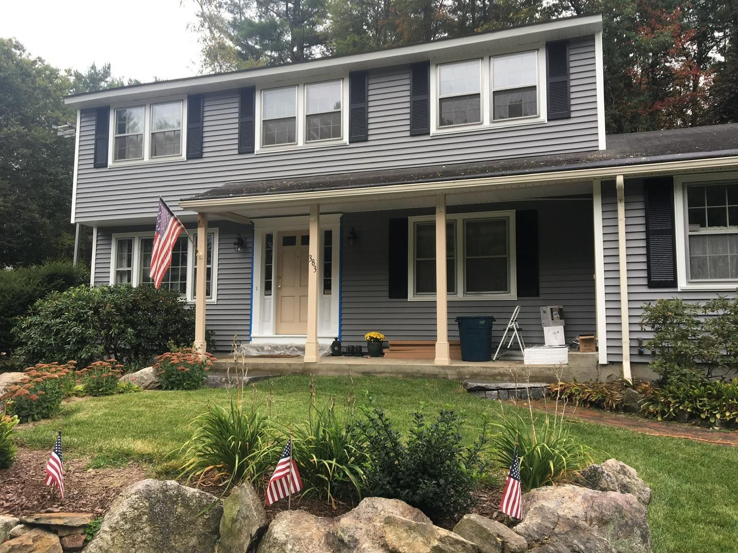 Two-story gray house with white trim, black shutters, American flag, and a porch.