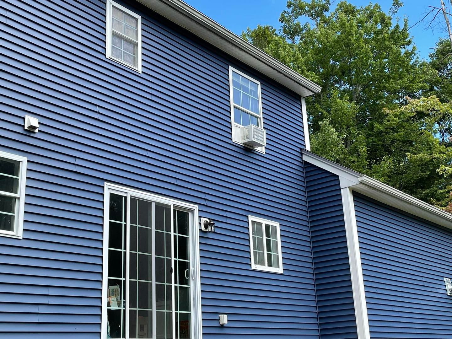 Blue-sided house with white-framed windows and sliding glass doors; a/c unit visible. Green trees in background.