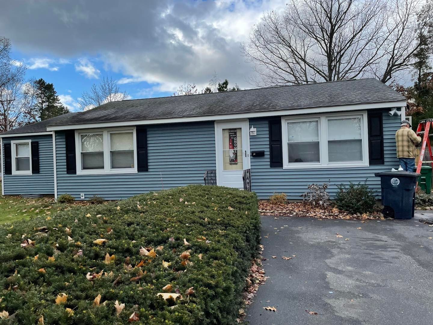 Blue house with black shutters, a trimmed bush in front, and a person on a ladder on the right.