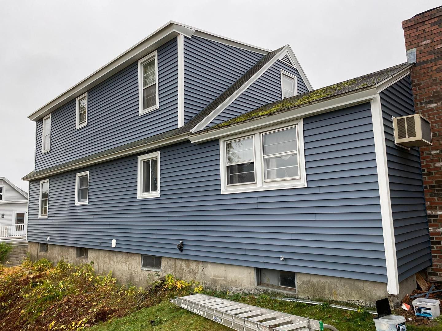 Blue-sided house with white trim. Air conditioner on brick chimney. Overcast day.