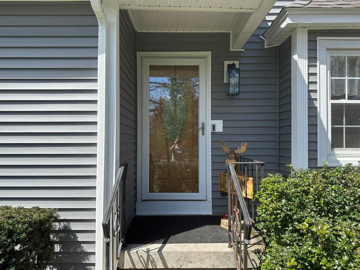 White-framed glass door on a gray house with a black welcome mat; metal railing and a decorative deer.