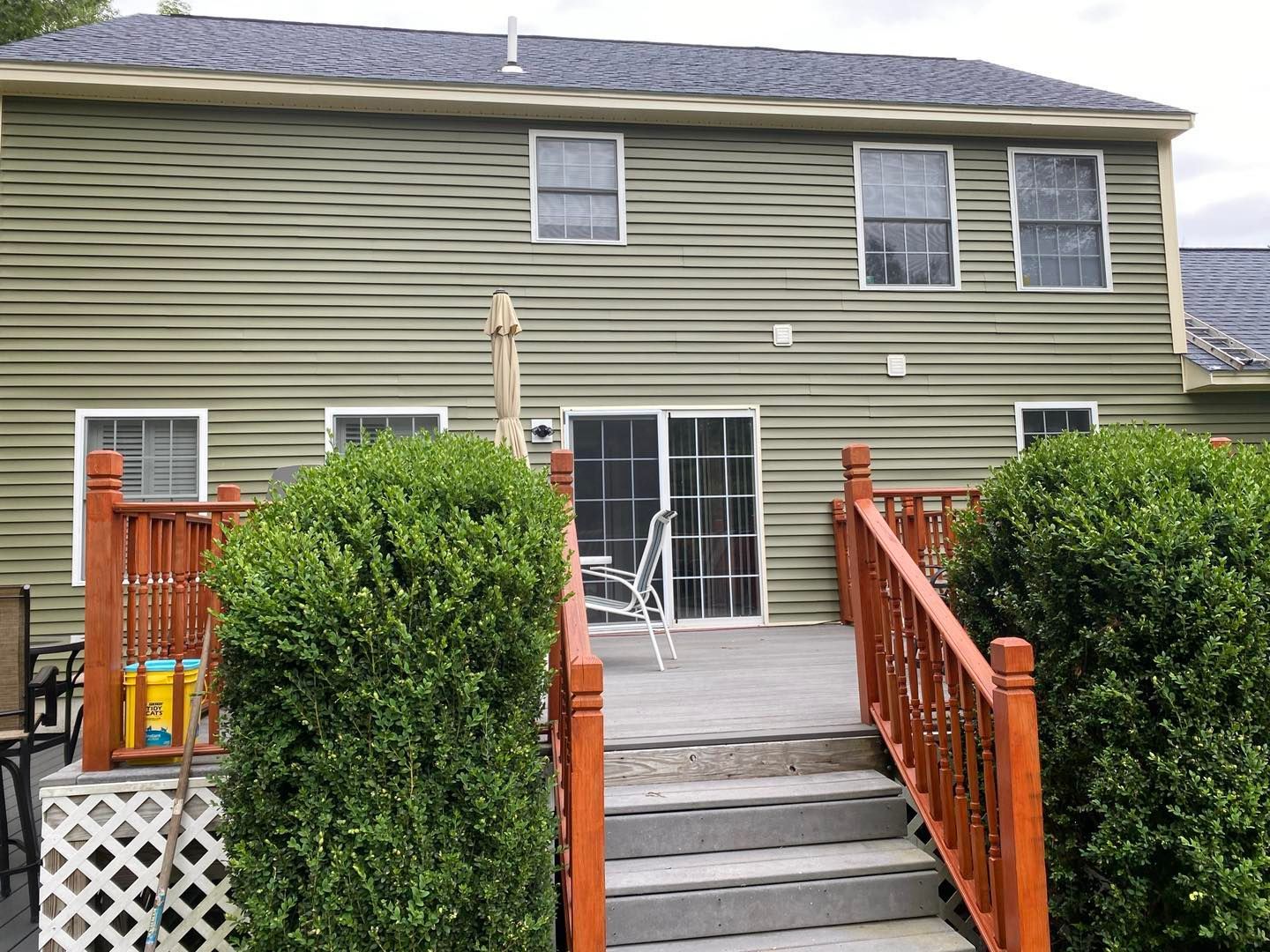 Green siding on a two-story house with a deck and steps leading up, surrounded by bushes.