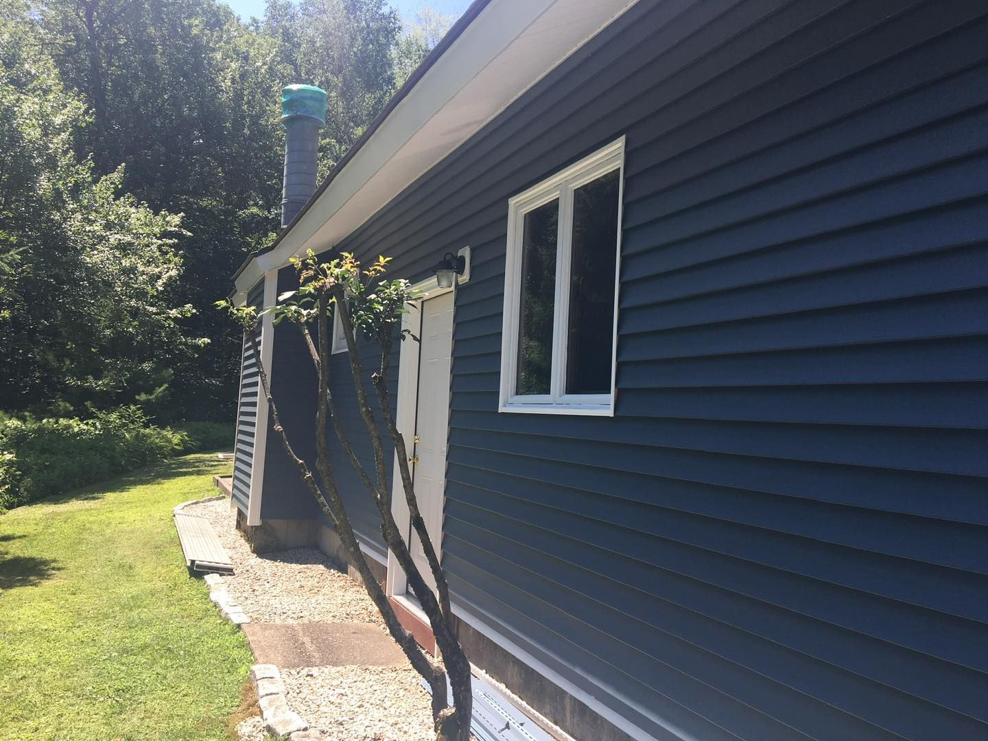 Blue-sided house with white window, door, and trim. Green grass and trees in the background.