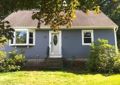 Blue-sided house with white trim, a bay window, and a front door, framed by green trees and bushes.
