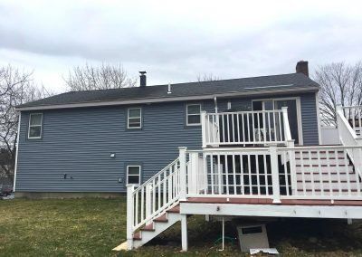 Two-story blue house with white deck and railing, on a grassy hill under a cloudy sky.