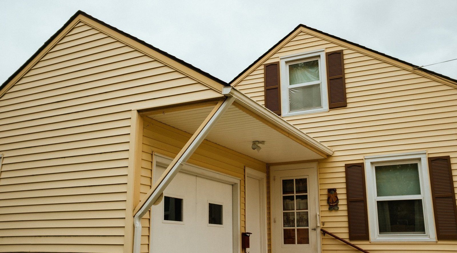 Yellow house with white garage door and brown shutters under a cloudy sky.