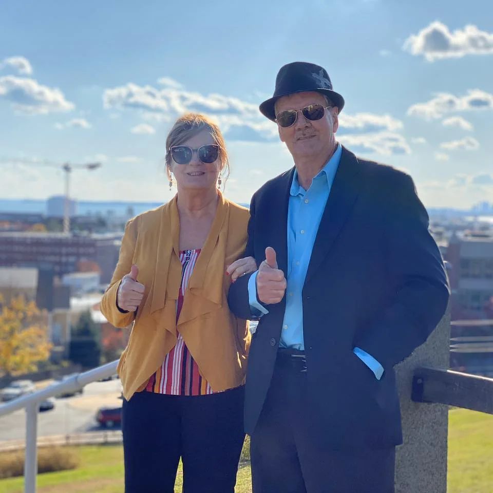 A couple smiles, giving thumbs up outdoors. The woman wears yellow, the man a black suit and hat. City in background.