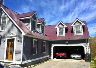 Gray house with a red metal roof, garage, and two cars under a blue sky.