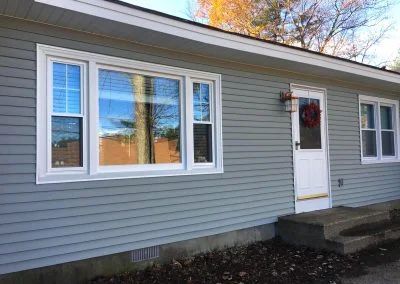 Blue-gray house with white trim windows and door; a red wreath hangs on the door, autumn leaves.