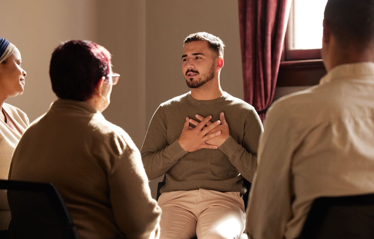 Group therapy session: Man speaking with hands on chest, surrounded by others in a circle, in a sunlit room.